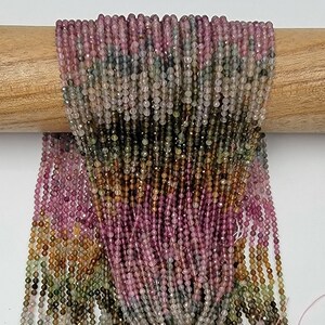 May include: Multiple strands of small, faceted, round beads in a gradient of colors including pink, green, brown, and translucent gray. The beads are strung together in long, vertical lines, creating a dense, textured, and colorful display.