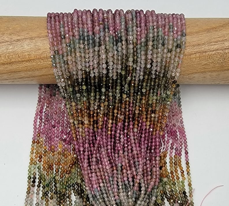 May include: Multiple strands of small, faceted, round beads in a gradient of colors including pink, green, brown, and translucent gray. The beads are strung together in long, vertical lines, creating a dense, textured, and colorful display.
