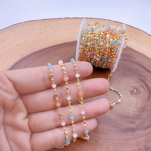 May include: A spool of gold-colored beaded chain with pastel-colored beads in shades of peach, pink, blue, and white. The chain is displayed on a wooden surface.