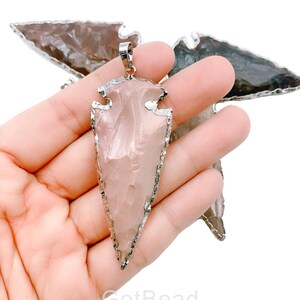 May include: A pink arrowhead pendant with a silver-toned metal edge and a silver-toned bail. The pendant is held in a hand.