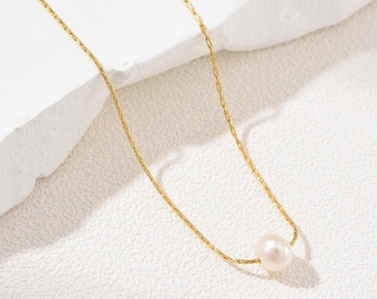May include: A delicate gold necklace featuring a single, lustrous pearl pendant. The chain has a fine, textured design, and the pearl is a soft, creamy white. The necklace is displayed against a white, textured backdrop.
