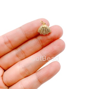 May include: A gold-colored seashell charm with a loop for attaching to a necklace or bracelet. The charm is encrusted with small, clear crystals, adding sparkle. The seashell design is detailed and elegant, suitable for jewelry making.