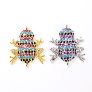 May include: Two frog-shaped pendants, one gold-toned and one silver-toned, each encrusted with colorful gemstones. The gemstones are arranged in a pattern of black, turquoise, and magenta. The pendants are on a white background.