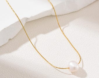 May include: A delicate gold-tone necklace featuring a single, luminous white pearl pendant. The chain has a fine, textured design. The pearl is smooth and round, creating a classic and elegant look. Jewelry.