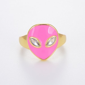 Puede incluir: Un anillo de oro con un diseño de cara de extraterrestre rosa y diamantes de imitación transparentes para los ojos.