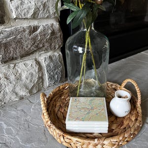 May include: A wicker tray with a stack of square coasters featuring a map design. A small white ceramic jar with the word "Pickles" on it is on the tray. A clear glass vase with olive branches is in the background.