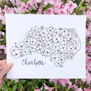 May include: A white print of the state of North Carolina with the city of Charlotte written below. The state is outlined in black and filled with white dogwood flowers.