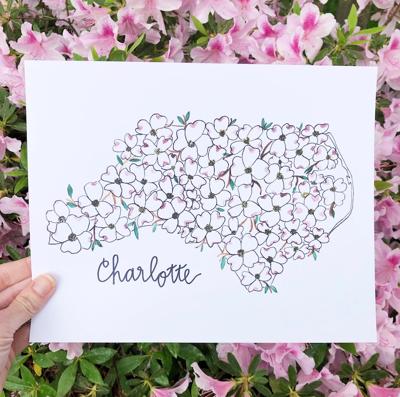 May include: A white print of the state of North Carolina with the city of Charlotte written below. The state is outlined in black and filled with white dogwood flowers.