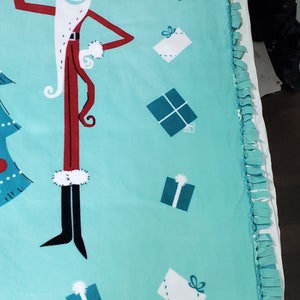 May include: A turquoise blue fleece blanket with a tie-dye fringe border. The blanket features a cartoon image of Jack Skellington dressed as Santa Claus with presents.