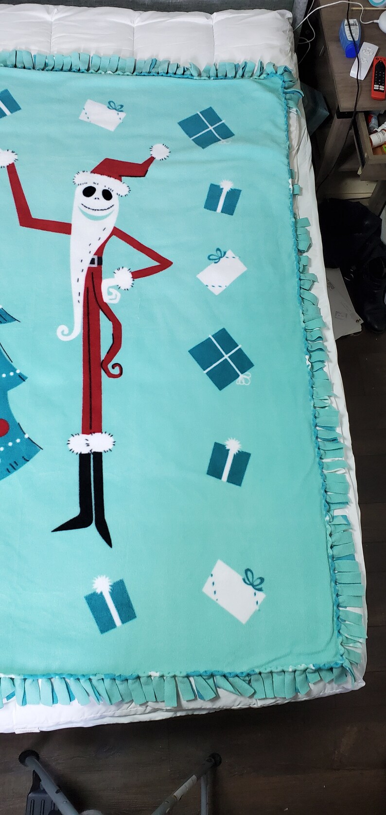 May include: A turquoise blue fleece blanket with a tie-dye fringe border. The blanket features a cartoon image of Jack Skellington dressed as Santa Claus with presents.