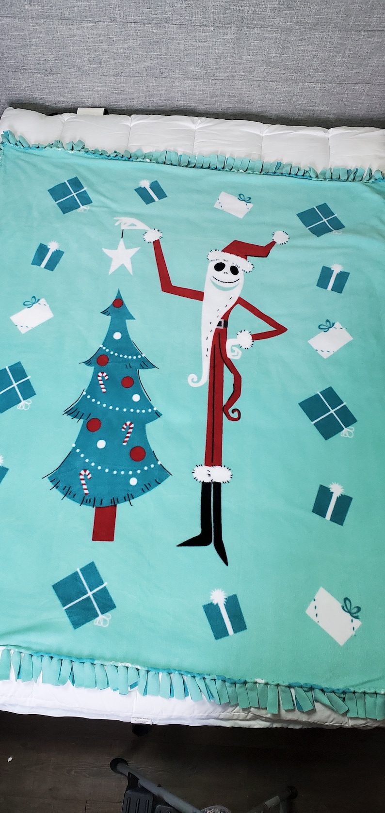 May include: A turquoise blue fleece blanket featuring a Christmas tree and a character dressed as Santa Claus. The character is Jack Skellington from the film The Nightmare Before Christmas. The blanket has a fringed border and is decorated with presents.