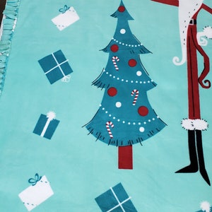 May include: A turquoise blue fleece tie blanket with a Christmas tree, presents, and a character resembling Jack Skellington from The Nightmare Before Christmas. The blanket has a fringe border.