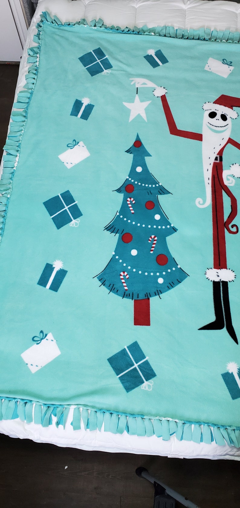 May include: A turquoise blue fleece tie blanket with a Christmas tree, presents, and a character resembling Jack Skellington from The Nightmare Before Christmas. The blanket has a fringe border.