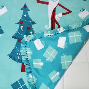 May include: A turquoise blue fleece blanket featuring a Christmas tree and a character from The Nightmare Before Christmas dressed as Santa Claus. The blanket has a fringed border and a pattern of white and turquoise blue gift boxes.