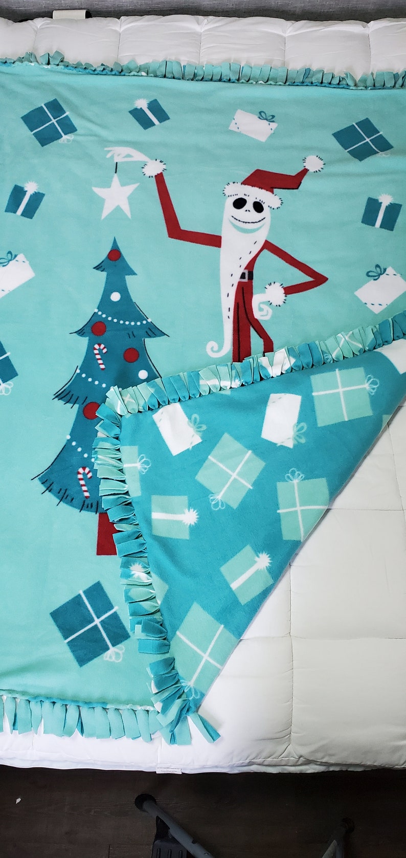 May include: A turquoise blue fleece blanket featuring a Christmas tree and a character from The Nightmare Before Christmas dressed as Santa Claus. The blanket has a fringed border and a pattern of white and turquoise blue gift boxes.