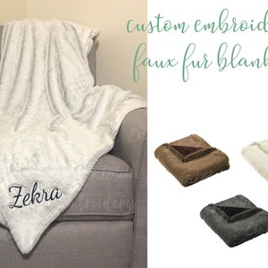 May include: A white faux fur blanket with the name "Zekra" embroidered on it. The blanket is folded and displayed on a gray armchair. Three additional faux fur blankets in brown, white, and gray are shown below the armchair.