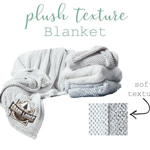 May include: A white plush blanket with a soft, textured weave. The blanket has a logo that says "Grand Lodge" with a pine tree graphic. The blanket is folded and stacked on a white surface.