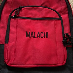 Op de afbeelding: Rode rugzak met zwarte accenten en een groot voorvak. Het vak heeft een zwarte rits en de naam "MALACHI" is in het zwart geborduurd op de voorkant.