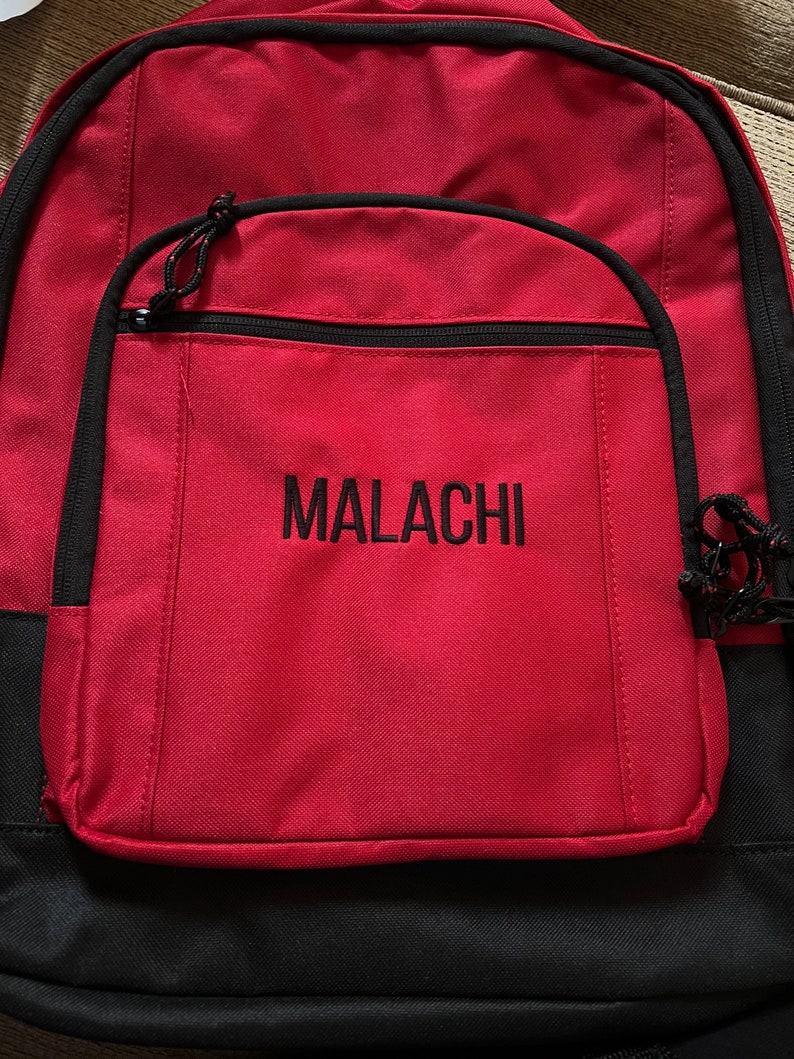 Op de afbeelding: Rode rugzak met zwarte accenten en een groot voorvak. Het vak heeft een zwarte rits en de naam "MALACHI" is in het zwart geborduurd op de voorkant.