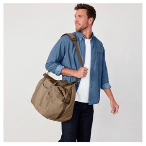 May include: A large olive green canvas duffel bag featuring a structured rectangular base, two top handles, and an adjustable shoulder strap with a padded section. The bag includes dark brown leather-like accents and side pockets for storage.
