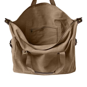 May include: A large, open-top tan canvas duffel bag featuring dark brown leather handle accents, a top zipper closure, and a single interior zippered pocket. The bag includes two sturdy fabric handles and side metal hardware for strap attachments.