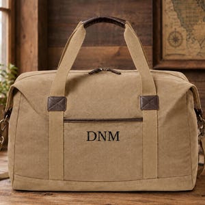 May include: A tan canvas duffel bag featuring dark brown leather accents on the handles and trim. The front of the bag displays the embroidered letters DNM in a dark font. It includes a detachable shoulder strap with metal hardware clips.