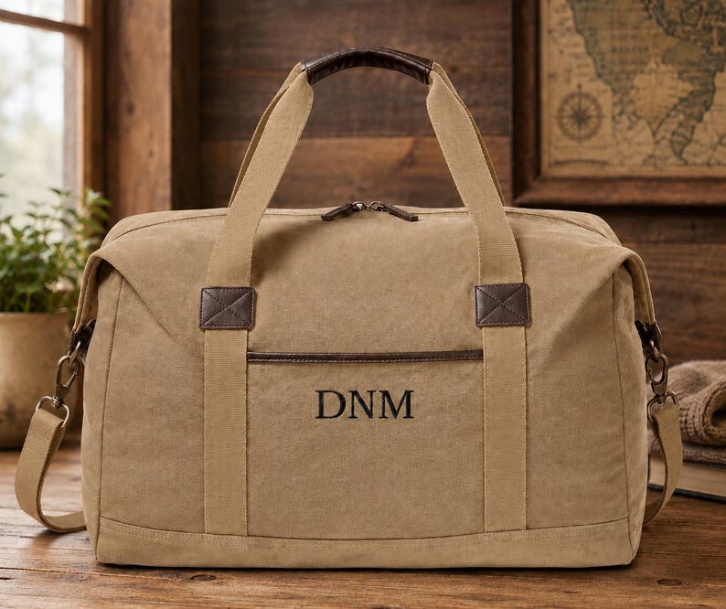 May include: A tan canvas duffel bag featuring dark brown leather accents on the handles and trim. The front of the bag displays the embroidered letters DNM in a dark font. It includes a detachable shoulder strap with metal hardware clips.