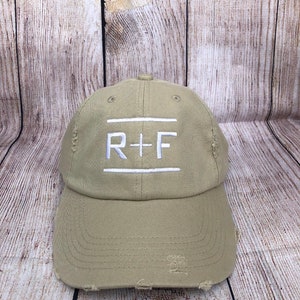 Może przedstawiać: Czapka bejsbolowa w kolorze khaki z przetartym wyglądem. Czapka ma białe haftowane logo z napisem "R+F" z przodu.