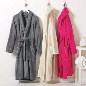 May include: Three bathrobes hanging on hooks. The bathrobes are gray, white, and pink. A white shelf with a white towel and a white vase is in the background.