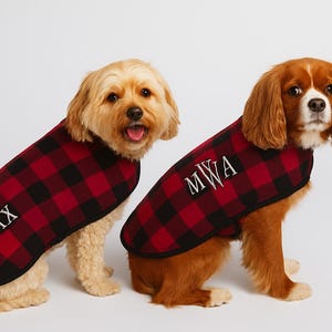 May include: Two dogs wearing matching red and black plaid dog coats. One coat has the name "MAX" embroidered in white, and the other has the initials "MWA". The dogs are sitting and looking at the camera.