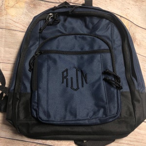 Op de afbeelding: Een donkerblauwe rugzak met een zwarte onderkant. De rugzak heeft een ritsvak aan de voorkant en een monogram van de letters "RUN" geborduurd in zwart.