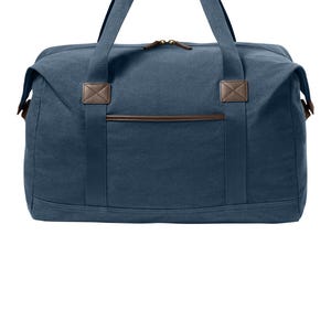 May include: A navy blue canvas duffel bag featuring brown faux leather accents on the handles, zipper pulls, and front pocket trim. The bag includes two top handles, a main zippered compartment, and a rectangular front pocket with stitched detailing.