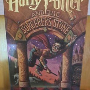 May include: A hardcover book titled Harry Potter and the Sorcerer's Stone by J.K. Rowling. The cover features a colorful illustration of a young person with glasses flying on a broomstick through a stone archway under a starry purple sky.