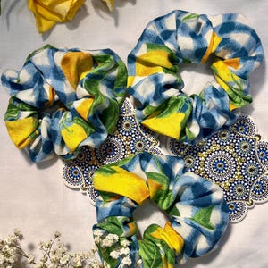 Op de afbeelding: Drie stoffen scrunchies met een citroen- en bladerenpatroon in geel, groen en blauw. De scrunchies zijn gerangschikt op een wit oppervlak met gele rozen en kleine witte bloemen. Decoratieve onderzetters zijn ook zichtbaar.