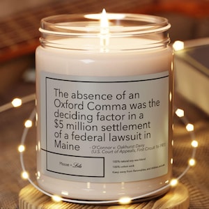 Peut inclure: Une bougie allumée dans un pot en verre transparent avec une étiquette blanche. L'étiquette indique : "L'absence d'une virgule d'Oxford a été le facteur décisif dans un règlement de 5 millions de dollars d'un procès fédéral dans le Maine." La bougie est sur un dessous de verre en bois.
