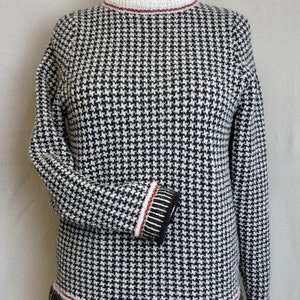 May include: A black and white houndstooth patterned sweater with a mock neck and red and black trim around the neck and hem.
