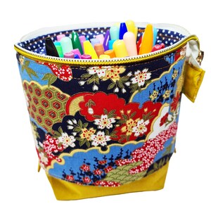 May include: A yellow fabric pencil holder with a zipper closure. The holder is decorated with a colorful Japanese-inspired floral pattern. The holder is filled with a variety of colored pens and pencils.