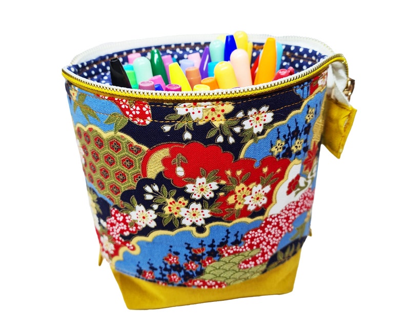 May include: A yellow fabric pencil holder with a zipper closure. The holder is decorated with a colorful Japanese-inspired floral pattern. The holder is filled with a variety of colored pens and pencils.