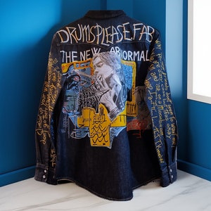 May include: Dark denim shirt with graffiti-style artwork. The back features a portrait and text that reads "DRUMS PLEASE FAB" and "THE NEW ABNORMAL". The sleeves and body are covered in additional handwritten phrases and drawings. A unique, statement piece.