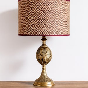 Op de afbeelding: Een tafellamp met een cilindrische lampenkap met een bruin en beige geweven ruitpatroon en een donkerrode rand. De voet is gemaakt van antiek goudkleurig metaal met een bolvormig, gestructureerd middenstuk en een geribbelde voet in voetstukstijl.