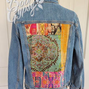 May include: Light blue denim jacket with a colorful patchwork design on the back. The patchwork features a variety of patterns and colors, including pink, green, orange, and purple. The jacket has a collar and button closures. The text "Boot Gypsy" is visible.