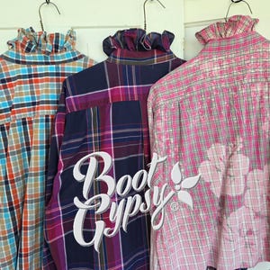 May include: Three long-sleeve button-down shirts with ruffled collars hanging on hangers. The left shirt has a blue, orange, and white plaid pattern. The middle shirt is dark purple with a plaid pattern. The right shirt is pink and white plaid with a floral design.