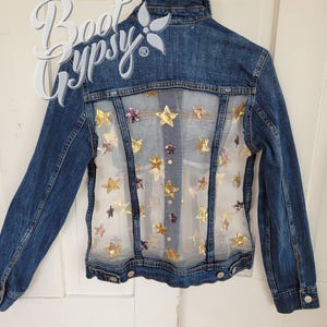 May include: A denim jacket with a sheer back panel adorned with gold and purple star-shaped sequins. The jacket has a classic collar, button closures, and long sleeves. The text "Boot Gypsy" is visible on the upper left.