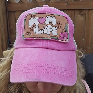 May include: Pink baseball cap with a mesh back and a curved brim. The cap features a patch with the words "MOM LIFE" in white bubble letters, a pink heart, and floral accents. The cap is a faded pink color.
