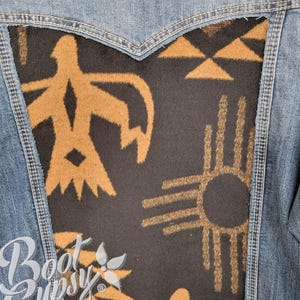 May include: A denim jacket with a black and tan patterned back panel. The tan design features geometric shapes and a sun symbol. The jacket has a classic denim wash and a standard collar.