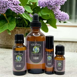 May include: Four bottles of essential oil with the label "Skylara Essentials Lilac" in various sizes. The bottles are on a light brown surface with a bouquet of purple lilac flowers in the background.