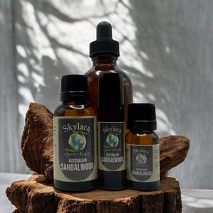 Puede incluir: Una colecci&oacute;n de botellas de aceite esencial y un frasco con cuentagotas, todos de vidrio &aacute;mbar, dispuestos sobre un trozo de madera. Las etiquetas dicen "Skylara essentials AUSTRALIAN SANDALWOOD". Las botellas var&iacute;an en tama&ntilde;o, con tapas negras y un aplicador de bola rodante.