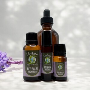 May include: Four amber glass bottles of essential oils with black caps. The bottles are labeled "Skylara essentials BEE BALM MONARDA". A dropper bottle, a roller bottle, and two smaller bottles are visible. A purple flower is in the background.