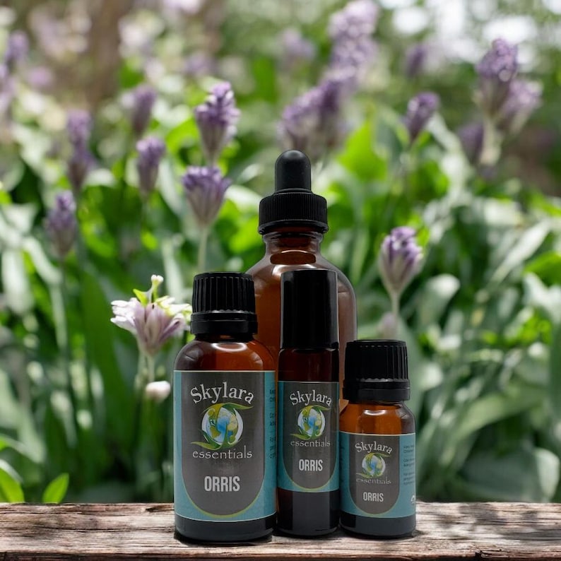 K&ouml;nnte beinhalten: Vier braune Glasflaschen mit &auml;therischen &Ouml;len, schwarzen Kappen und Etiketten mit der Aufschrift "Skylara essentials ORRIS". Der Hintergrund zeigt einen verschwommenen Garten mit lila Blumen und gr&uuml;nem Laub.
