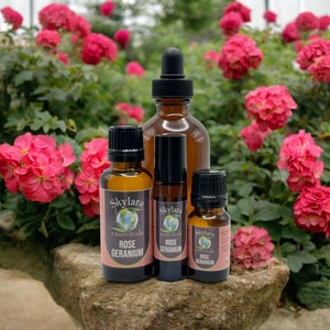May include: Four bottles of essential oil, labeled "Skylara Essentials Rose Geranium", are arranged on a rock with a background of pink roses.
